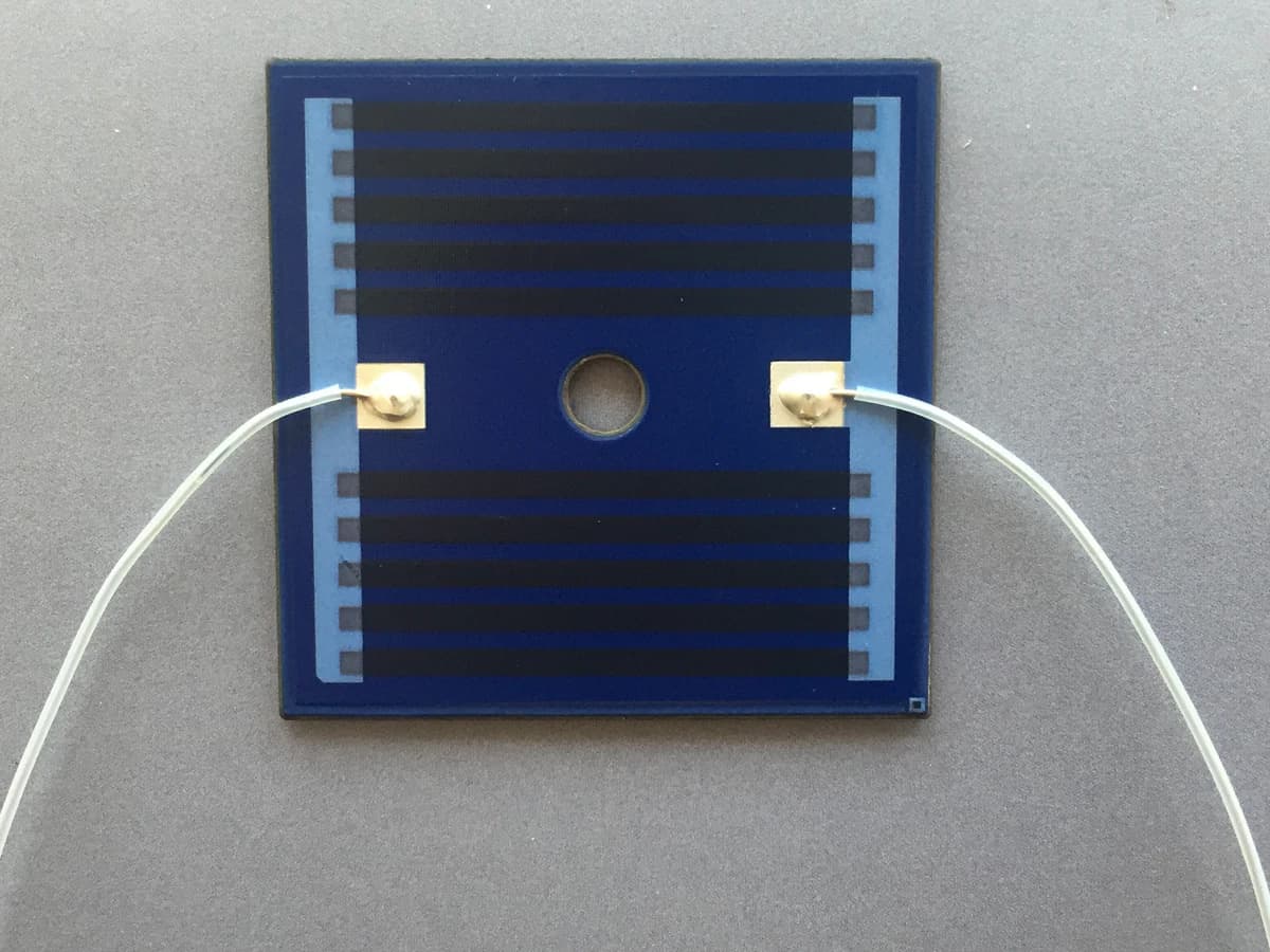 Square blue heater-product surface with dark printed tracks, a center opening, terminal pads, and white leads