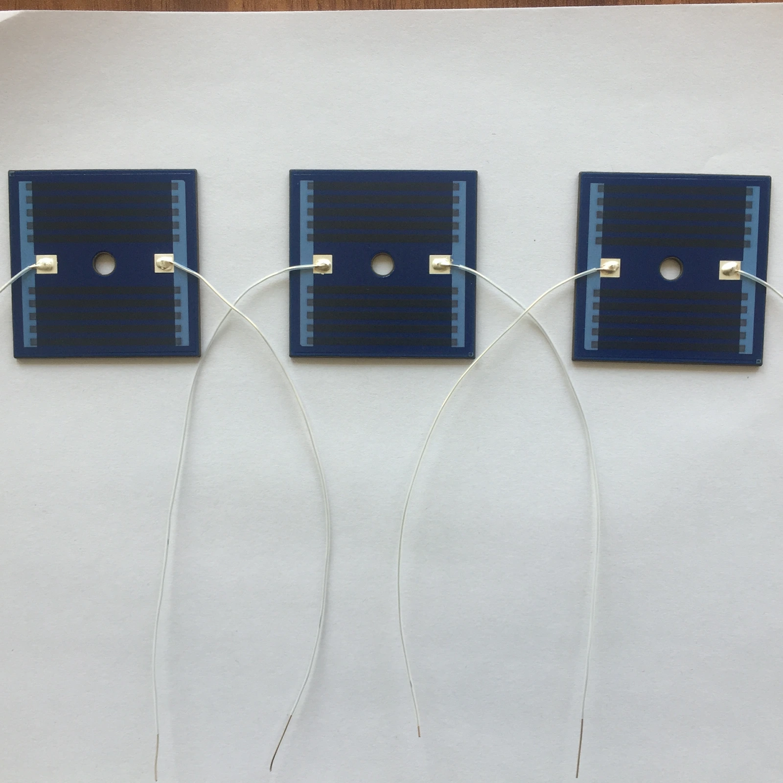 Three square heater plates with blue protective coating, central openings and attached white leads.