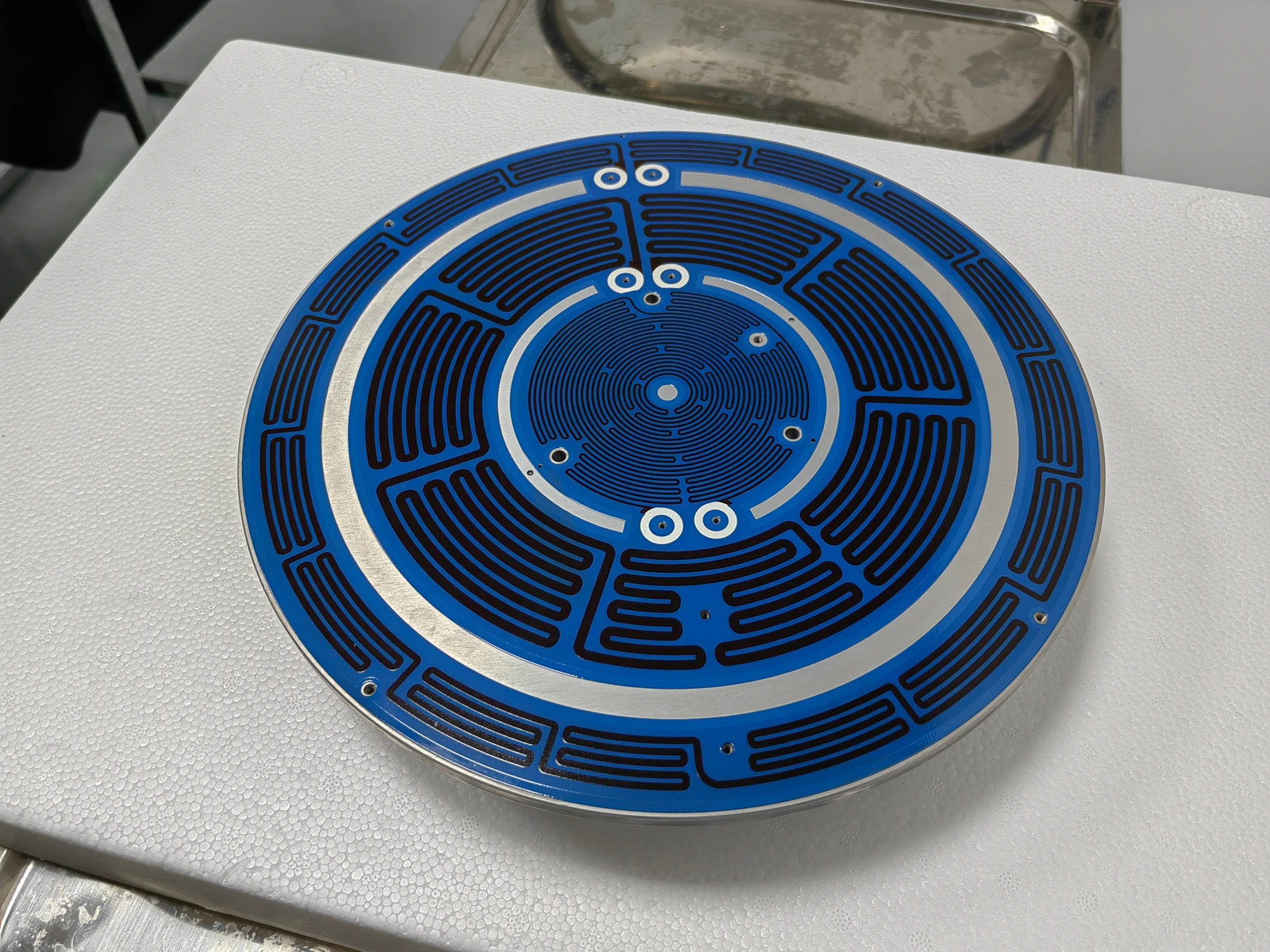 An oblique view of a circular blue-coated heater plate with patterned resistive paths and an exposed outer perimeter.