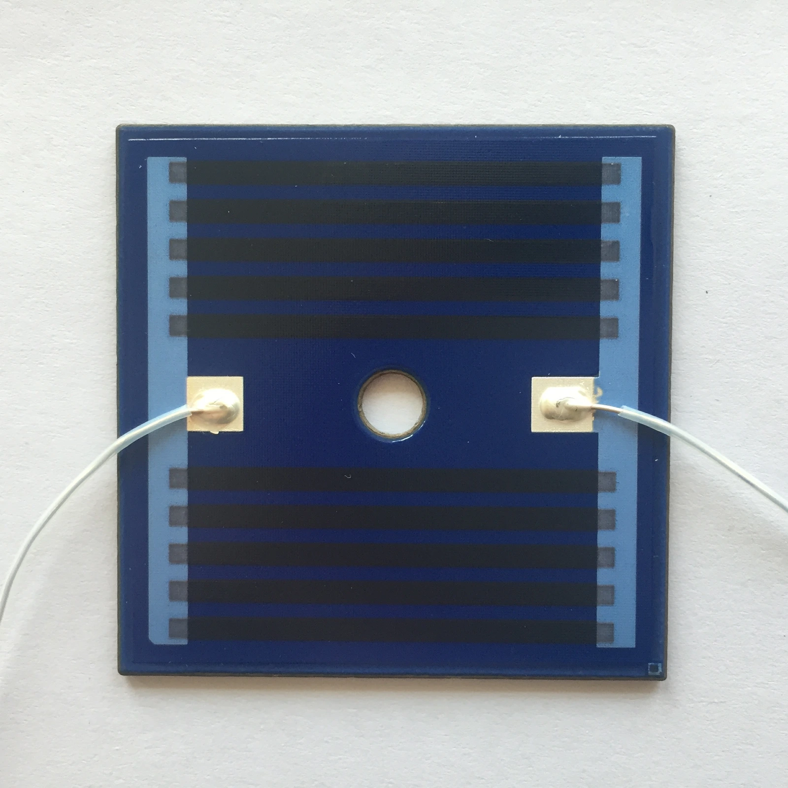 A square blue-coated heater with a central opening, parallel resistive paths and two white leads.