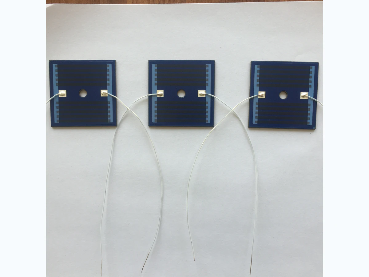 Three square stainless steel thick film heating plates with blue printed heater areas, center holes, terminals, and white leads